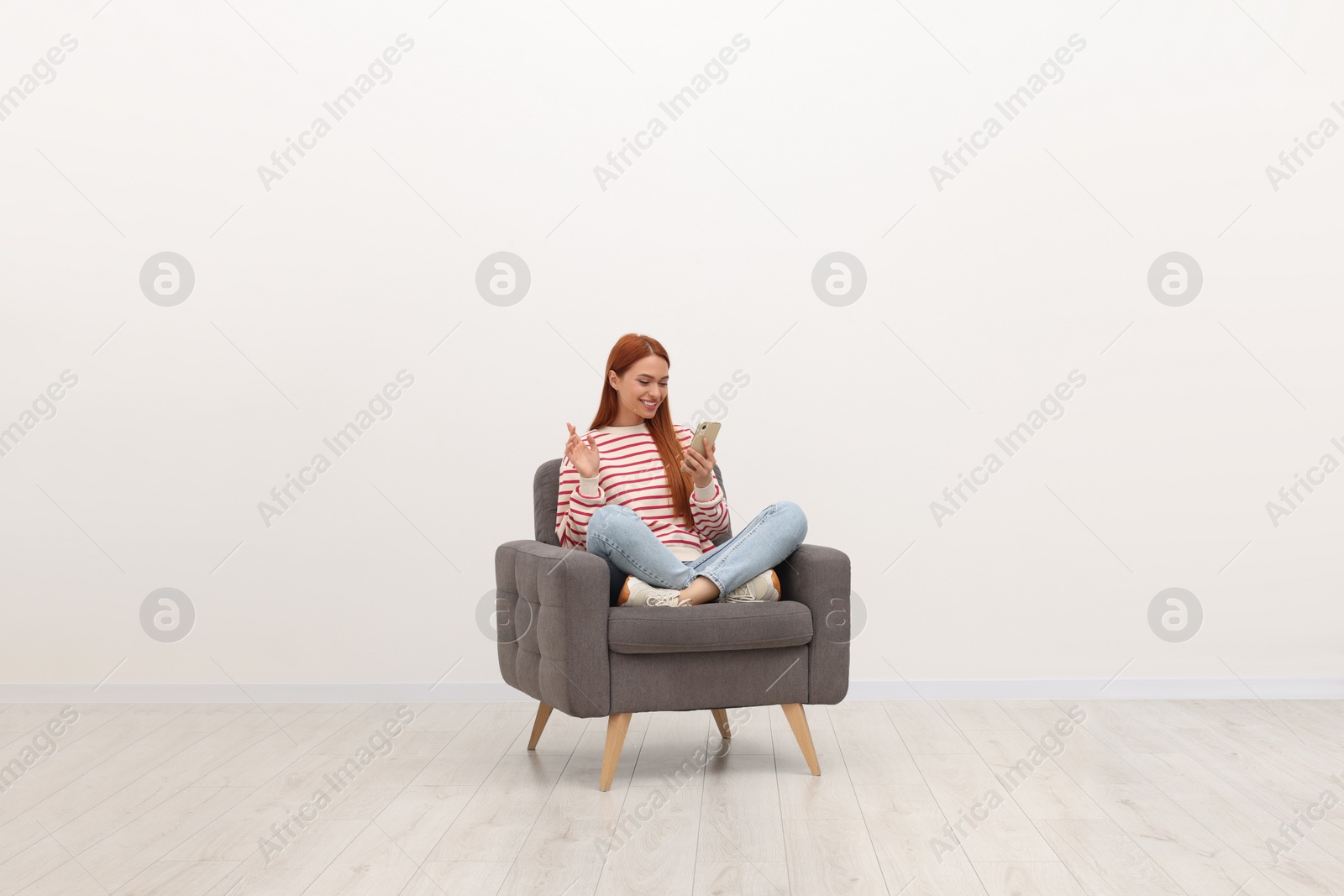 Photo of Happy young woman having video chat via smartphone while sitting in armchair near white wall indoors