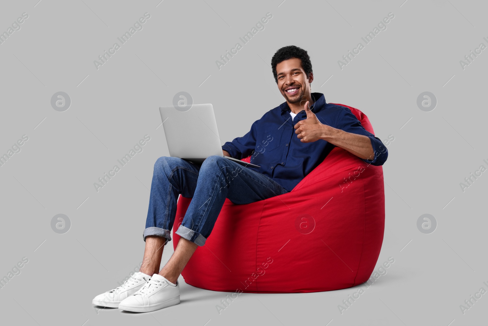 Photo of Happy man with laptop sitting on beanbag chair and showing thumb up against grey background