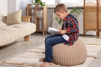 Photo of Boy with poor posture reading book on beige pouf in living room. Symptom of scoliosis