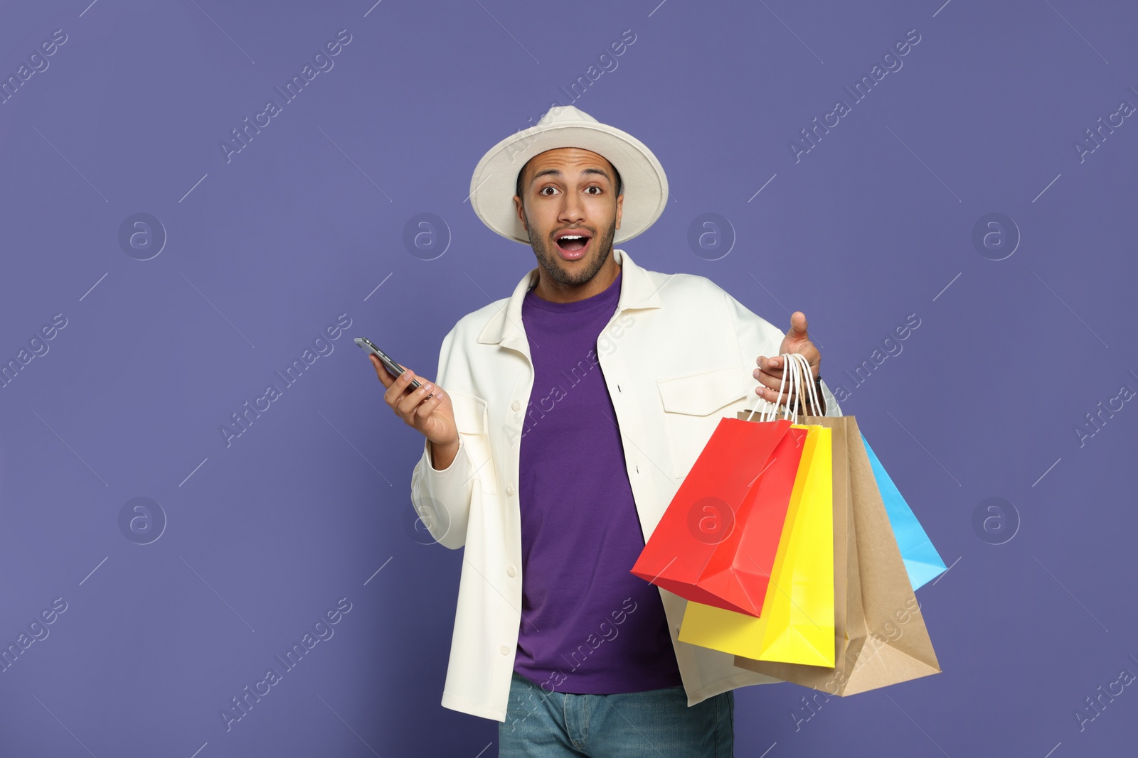 Photo of Emotional African American man in hat with shopping bags and smartphone on purple background