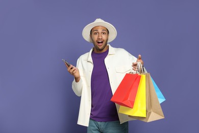 Photo of Emotional African American man in hat with shopping bags and smartphone on purple background
