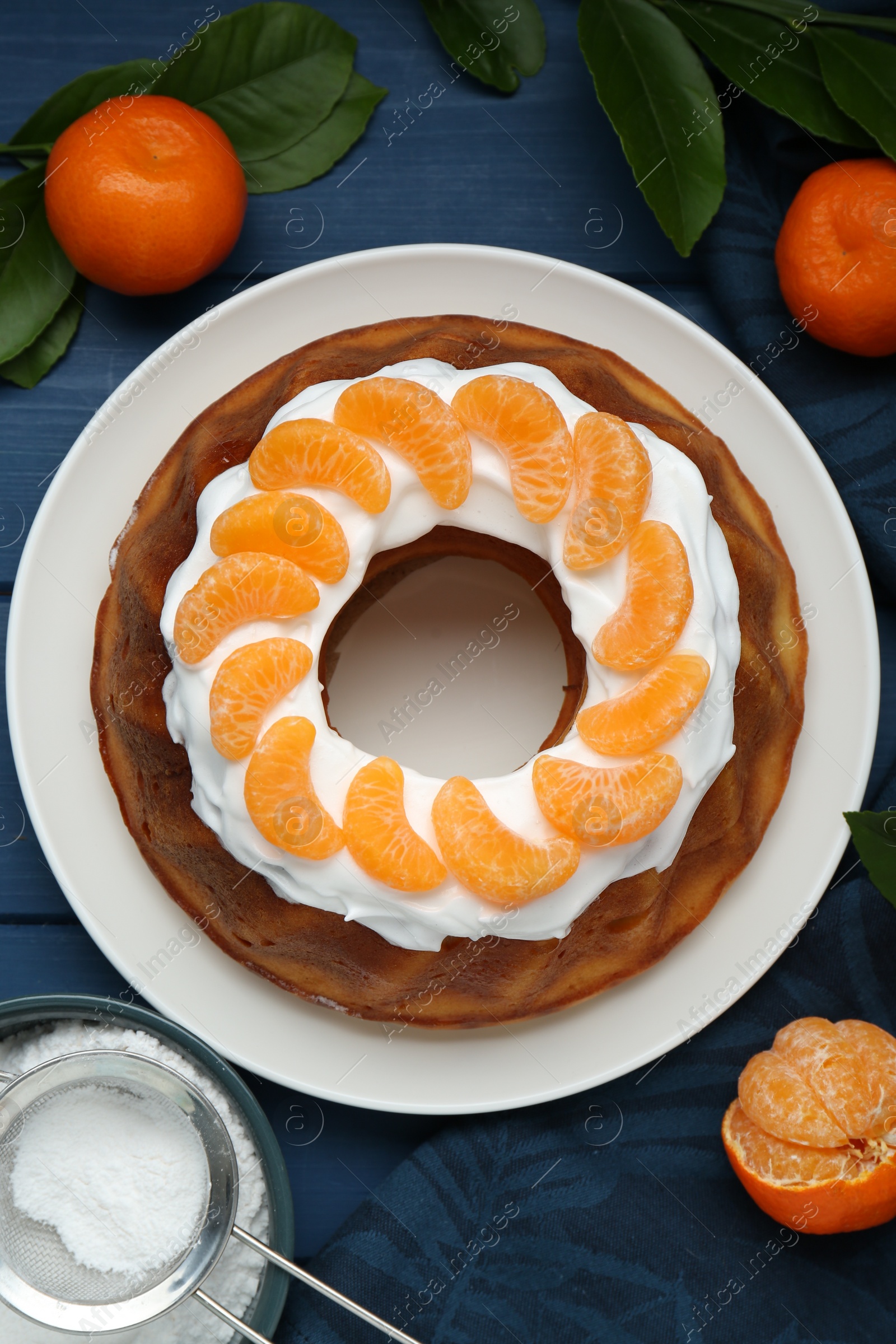 Photo of Homemade yogurt cake with tangerines, cream and green leaves on blue wooden table, flat lay