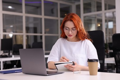 Photo of Woman taking notes while working near laptop at white desk in office