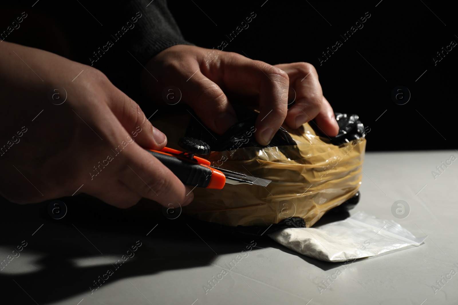 Photo of Smuggling and drug trafficking. Man opening package of narcotics with box cutter at light table against black background, closeup