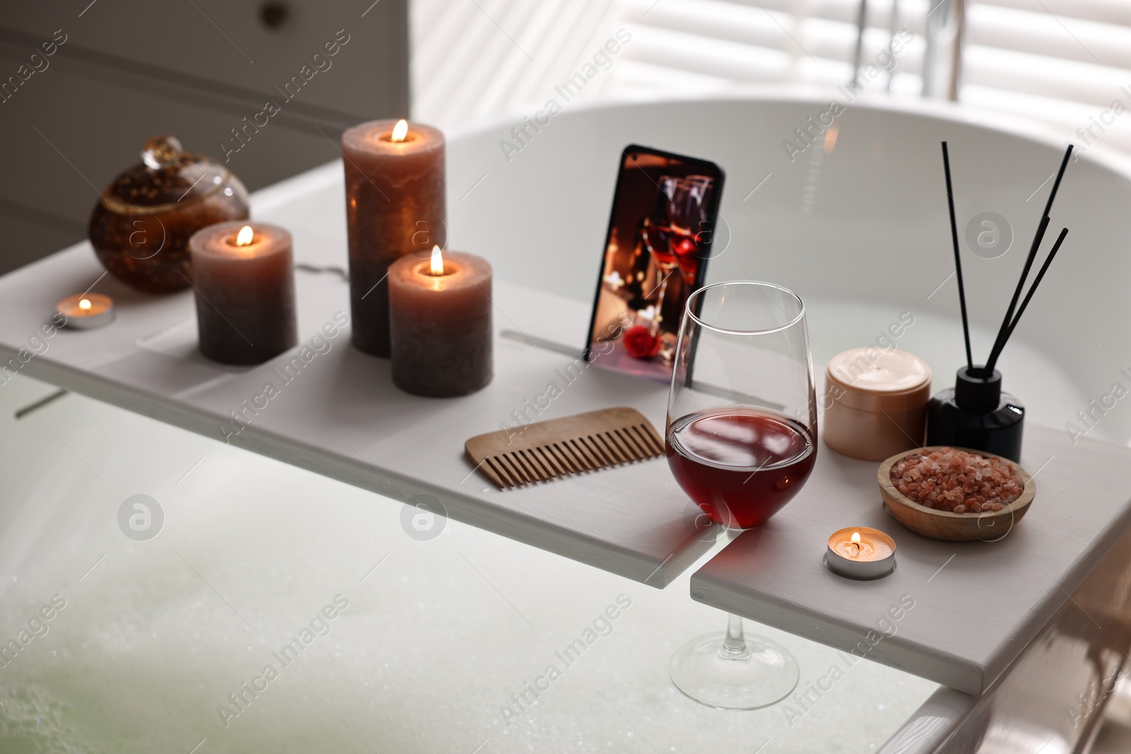 Photo of White wooden tray with smartphone, glass of wine and burning candles on bathtub in bathroom