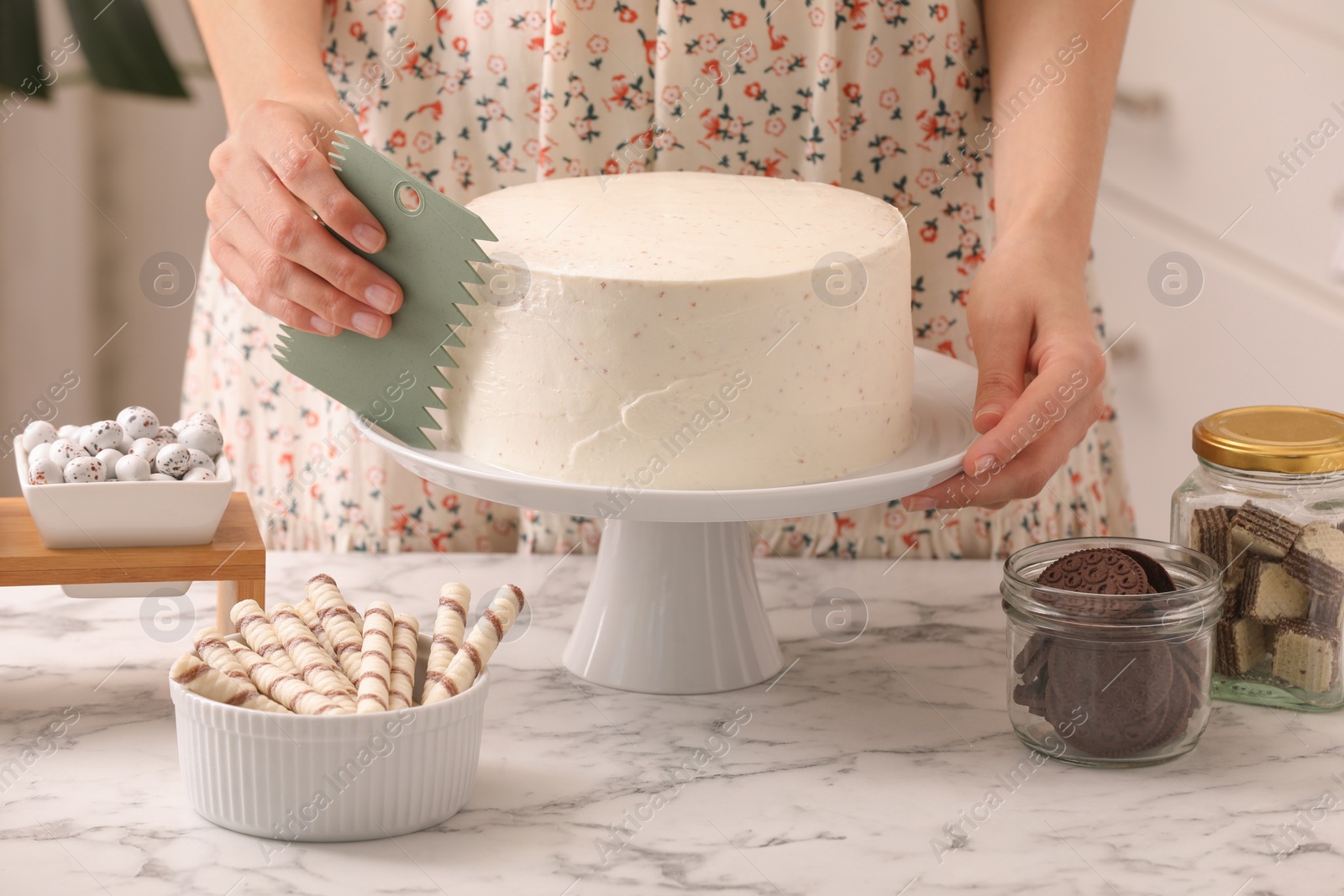 Photo of Woman using scraper to decorate cake at white marble table in kitchen, closeup