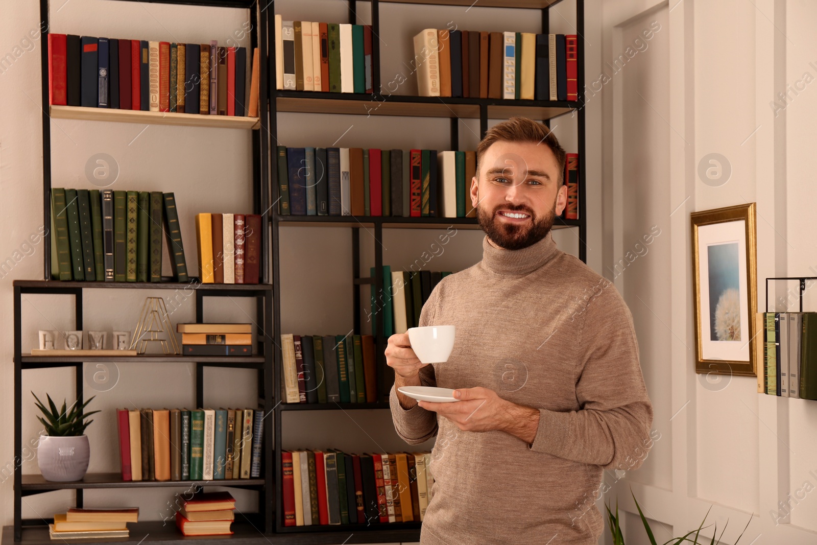 Photo of Young man with cup of coffee near shelves in home library