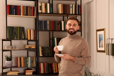 Young man with cup of coffee near shelves in home library