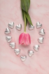 Heart made with delicious chocolate candies and beautiful tulip on pink table, flat lay