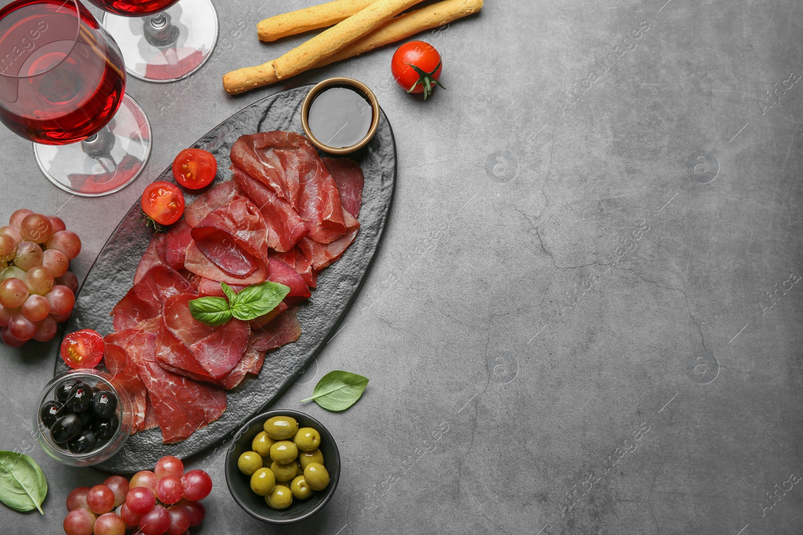 Photo of Delicious bresaola, tomato, olives, wine and basil leaves served on grey textured table, flat lay. Space for text