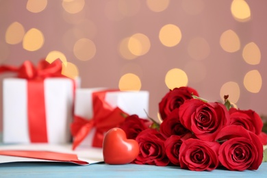 Photo of Beautiful red roses, decorative heart, love letter and gift boxes on table against blurred lights, space for text. St. Valentine's day celebration