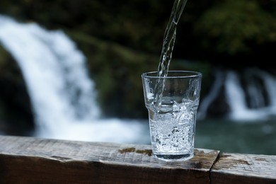 Photo of Fresh water pouring into glass on wooden surface near waterfall. Space for text