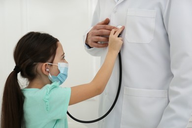 Photo of Little girl playing with pediatrician during visit in hospital. Patient wearing protective mask