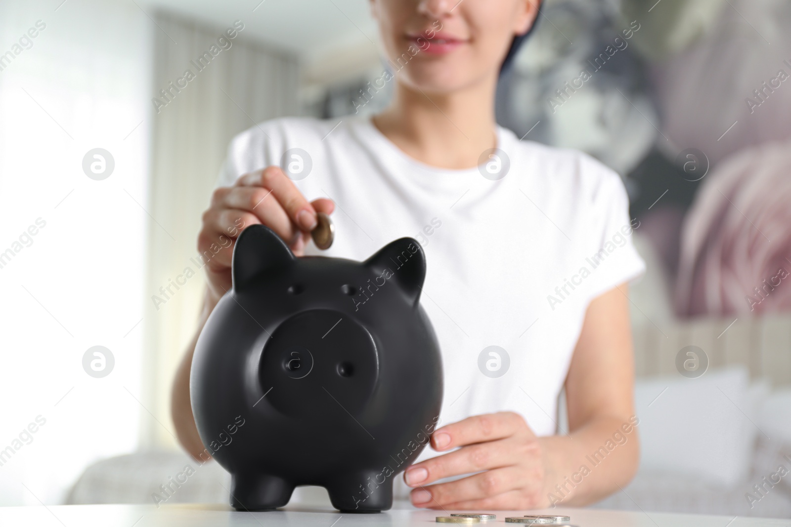 Photo of Woman putting money into piggy bank at white table indoors, closeup