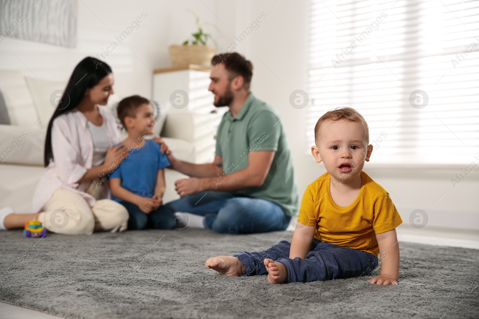 Photo of Unhappy baby sitting alone on floor while parents spending time with his elder brother at home. Jealousy in family