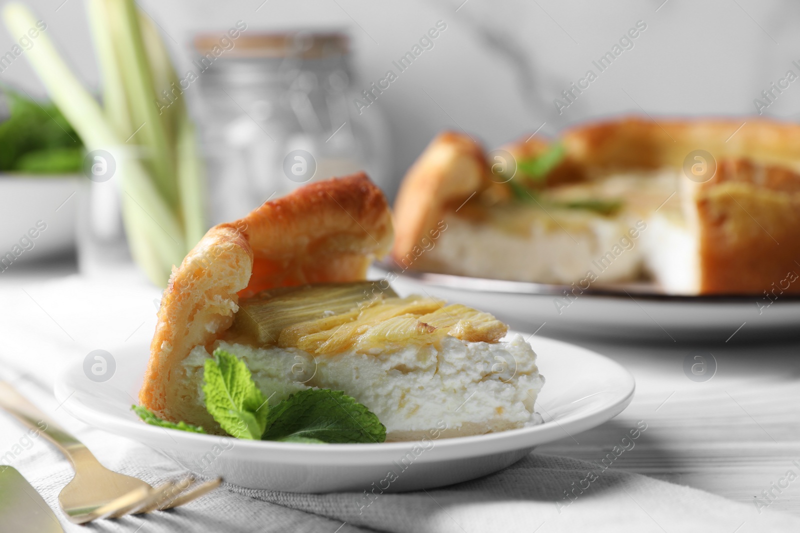 Photo of Piece of freshly baked rhubarb pie with cream cheese, mint leaves and cutlery on white wooden table, closeup