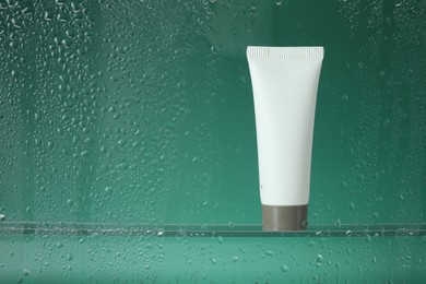 Tube with moisturizing cream on green background, view through wet glass. Space for text