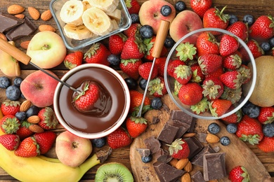 Photo of Fondue fork with strawberry in bowl of melted chocolate surrounded by other fruits on wooden table, flat lay