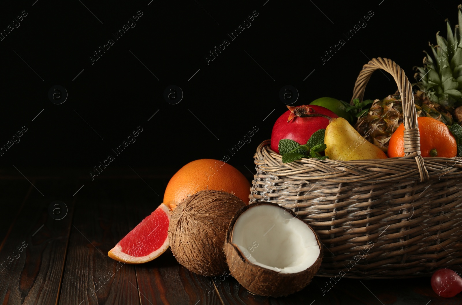 Photo of Wicker basket and different ripe fruits on wooden table against black background. Space for text