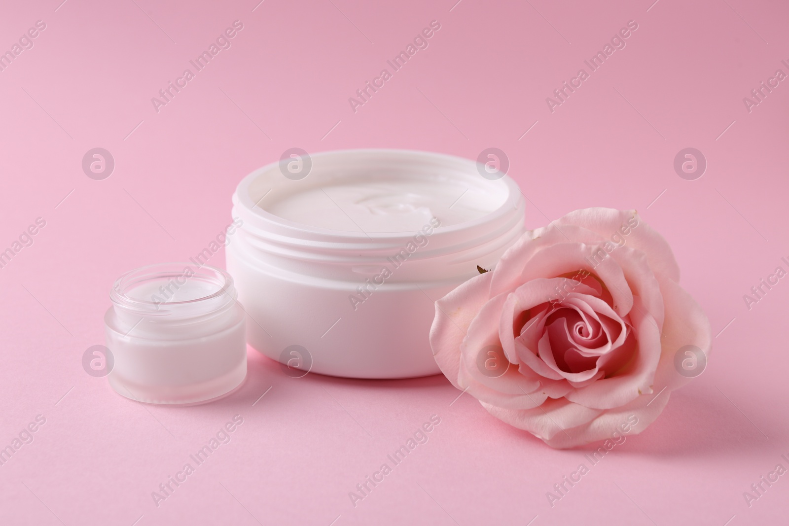 Photo of Moisturizing cream in open jars and rose flower on pink background. Body care product