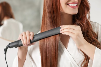 Photo of Young woman using hair iron indoors, closeup