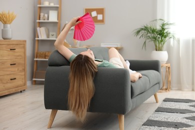Woman waving pink hand fan to cool herself on sofa at home