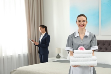 Photo of Chambermaid with stack of fresh towels and blurred supervisor in hotel room. Space for text