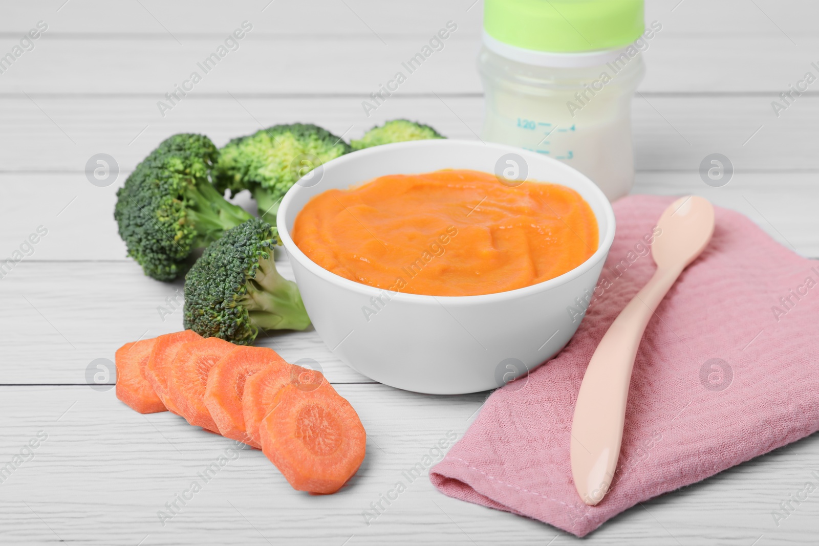 Photo of Bowl with healthy baby food, spoon and ingredients on white wooden table