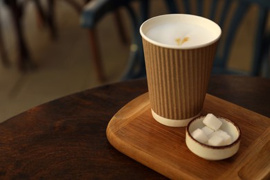 Photo of Delicious coffee in cardboard takeaway cup and bowl of sugar cubes on wooden table indoors, space for text