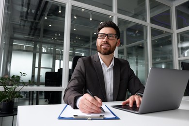 Photo of Man writing notes while working on laptop at white desk in office