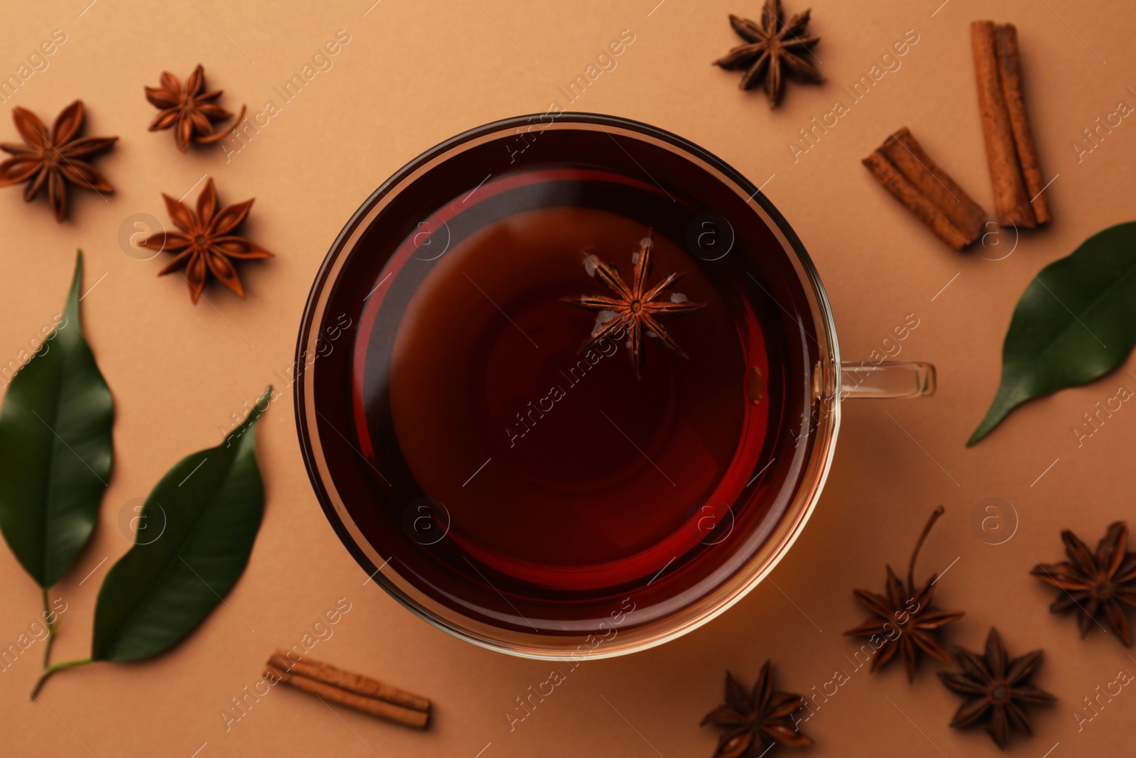 Photo of Cup of tea, anise stars, green leaves and cinnamon sticks on brown background, flat lay