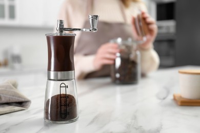 Photo of Woman opening glass jar with beans at table indoors, focus on coffee grinder. Space for text