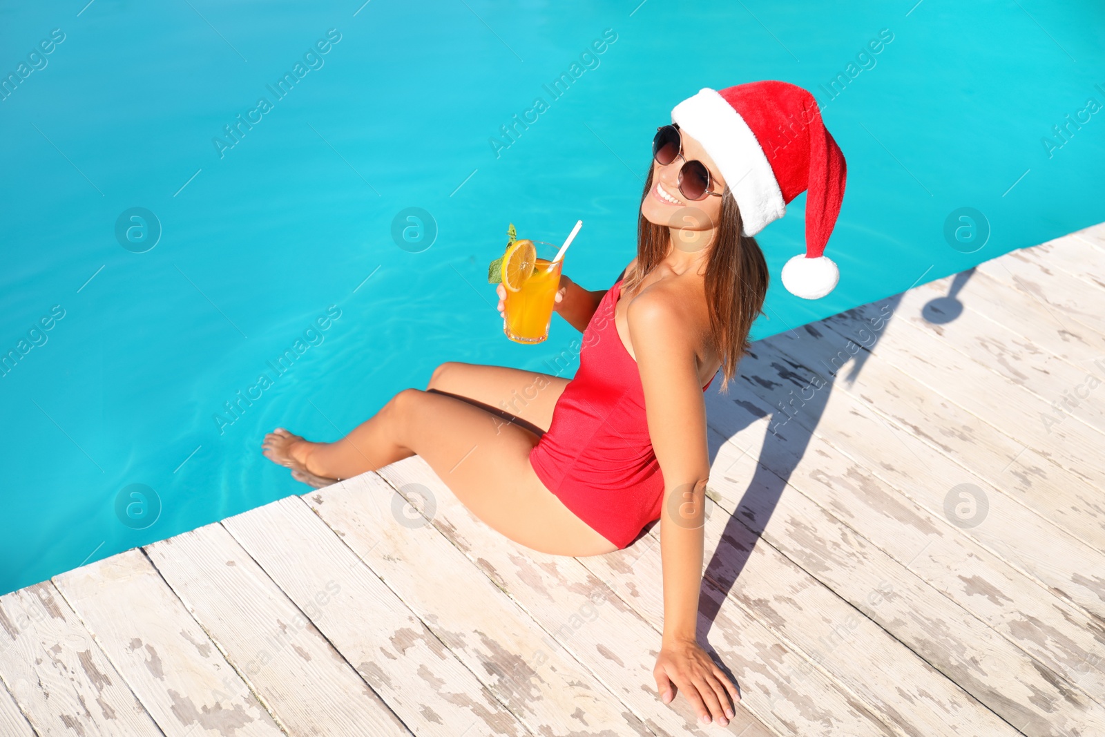 Photo of Young woman in Santa Claus hat with refreshing drink near swimming pool. Christmas vacation