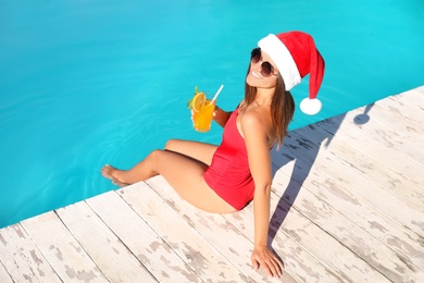 Photo of Young woman in Santa Claus hat with refreshing drink near swimming pool. Christmas vacation