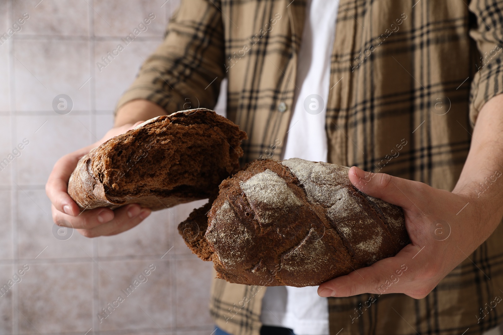 Photo of Man breaking loaf of fresh bread near grey wall, closeup