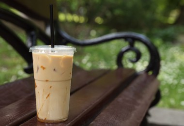 Takeaway plastic cup with cold coffee drink and straw on wooden bench outdoors, space for text