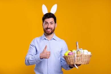 Portrait of happy man in cute bunny ears headband holding wicker basket with Easter eggs and showing thumb up on orange background