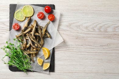 Slate board with delicious fried anchovies, lime and tomatoes on white wooden table, top view. Space for text