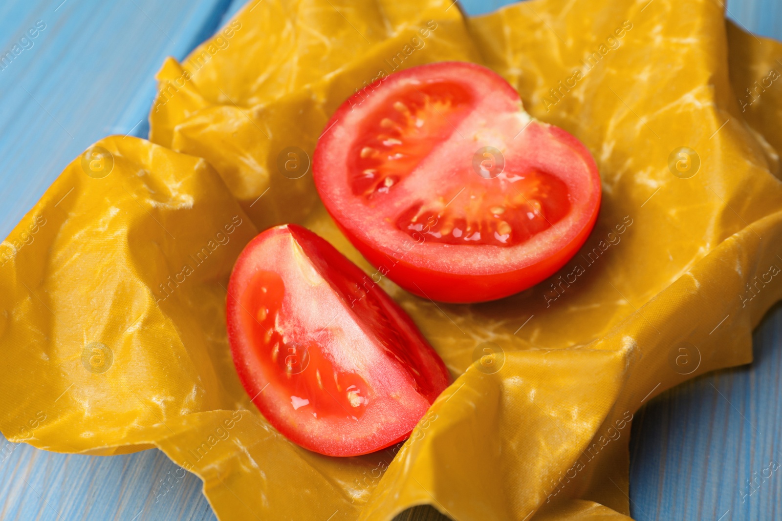 Photo of Slices of fresh tomato in beeswax food wrap on light blue wooden table, closeup