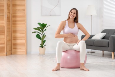 Pregnant woman sitting on fitness ball at home, space for text. Doing yoga