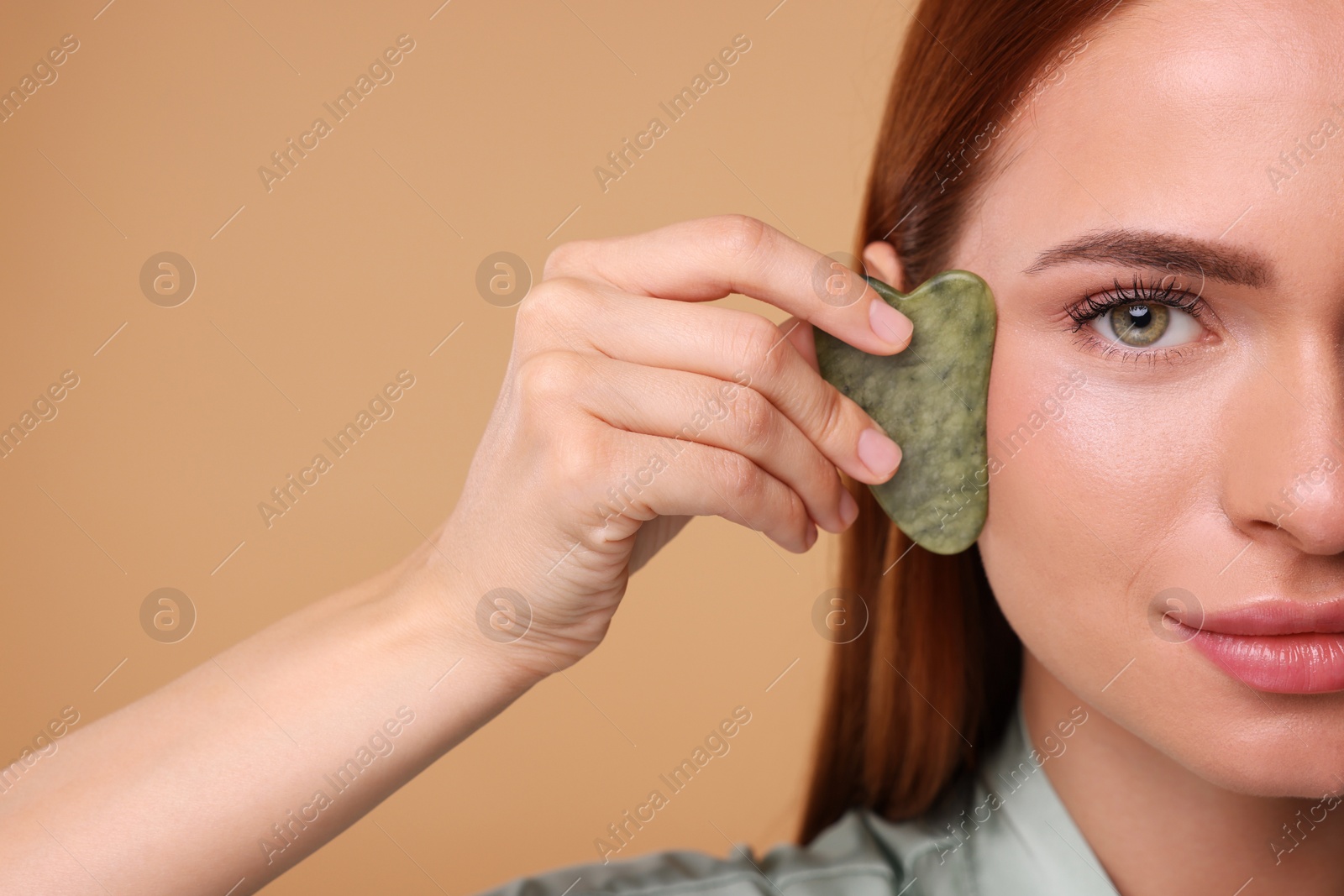 Photo of Young woman massaging her face with jade gua sha tool on pale orange background, closeup. Space for text
