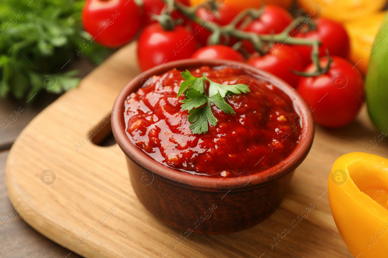 Photo of Delicious adjika sauce with parsley in bowl and ingredients on wooden board, closeup