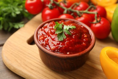 Photo of Delicious adjika sauce with parsley in bowl and ingredients on wooden board, closeup