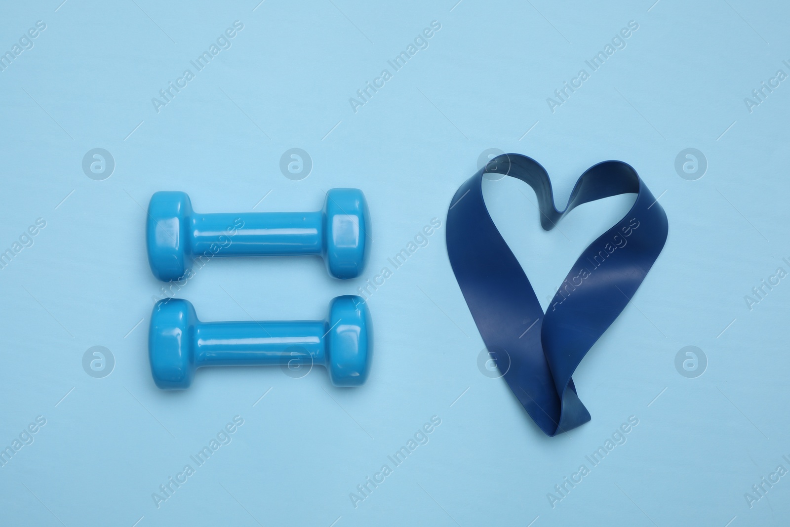 Photo of Two dumbbells and fitness elastic band in shape of heart on light blue background, flat lay. Lovely workout