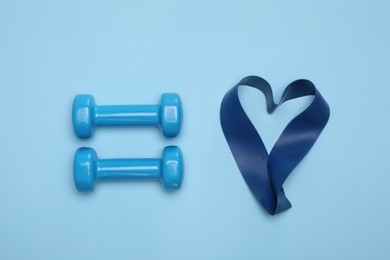 Two dumbbells and fitness elastic band in shape of heart on light blue background, flat lay. Lovely workout