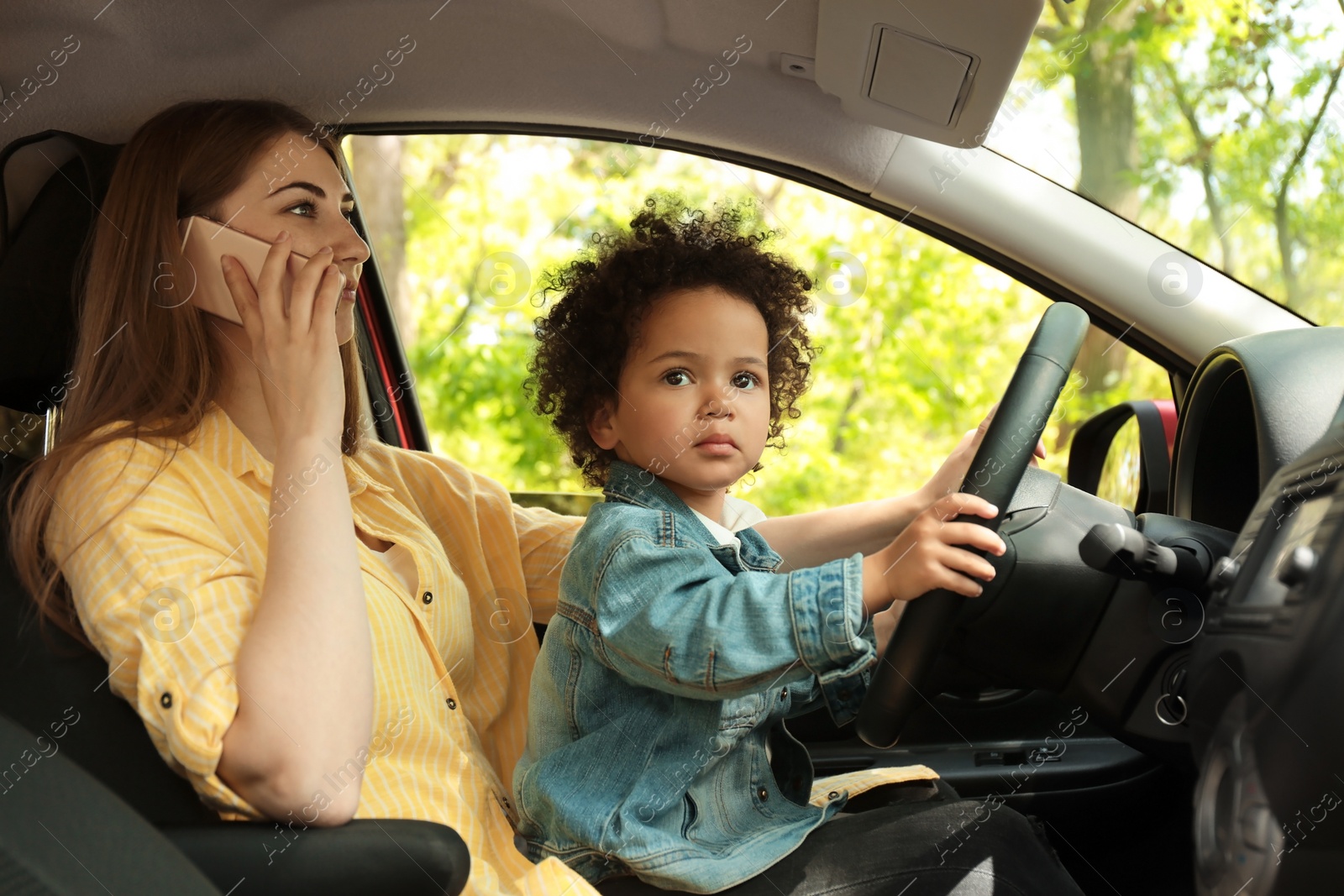 Photo of Mother with little daughter on knees driving car and talking by phone. Child in danger
