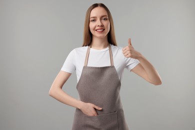 Photo of Beautiful young woman in kitchen apron showing thumbs up on grey background. Mockup for design