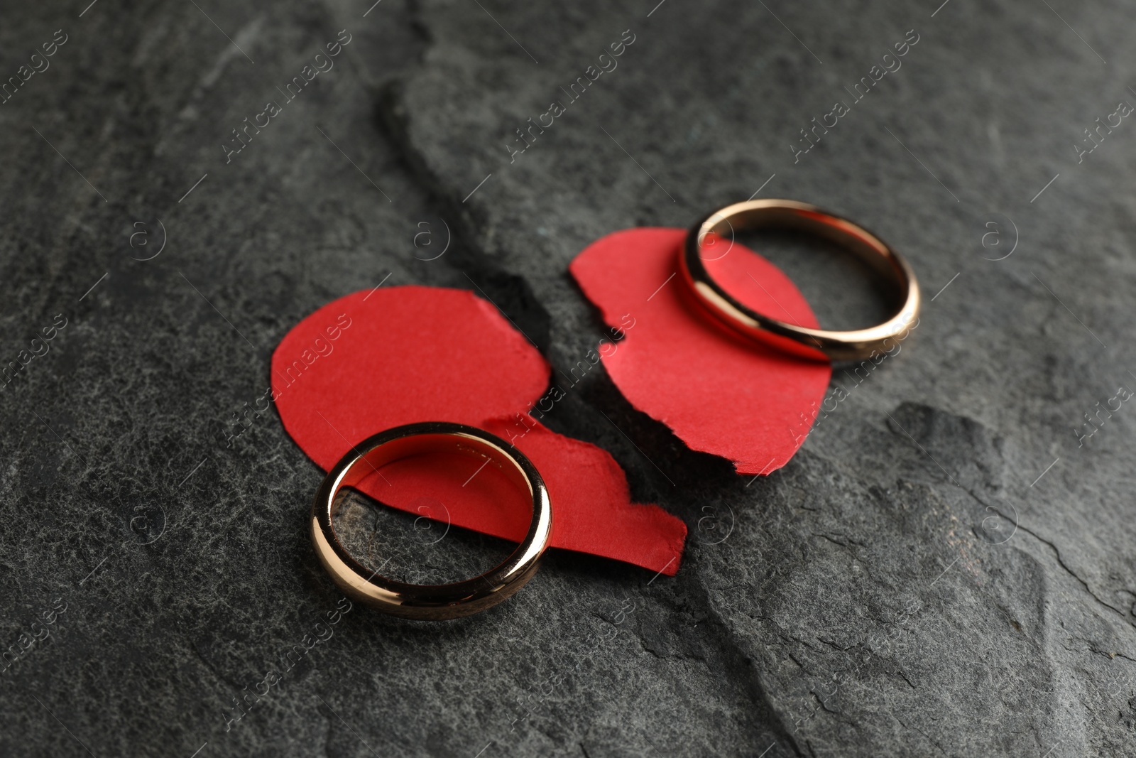 Photo of Halves of torn red paper heart and wedding rings on dark grey table. Broken heart