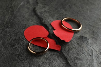 Halves of torn red paper heart and wedding rings on dark grey table. Broken heart