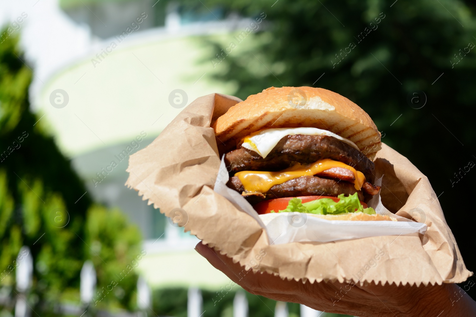 Photo of Woman holding delicious burger in paper wrap outdoors, closeup. Space for text